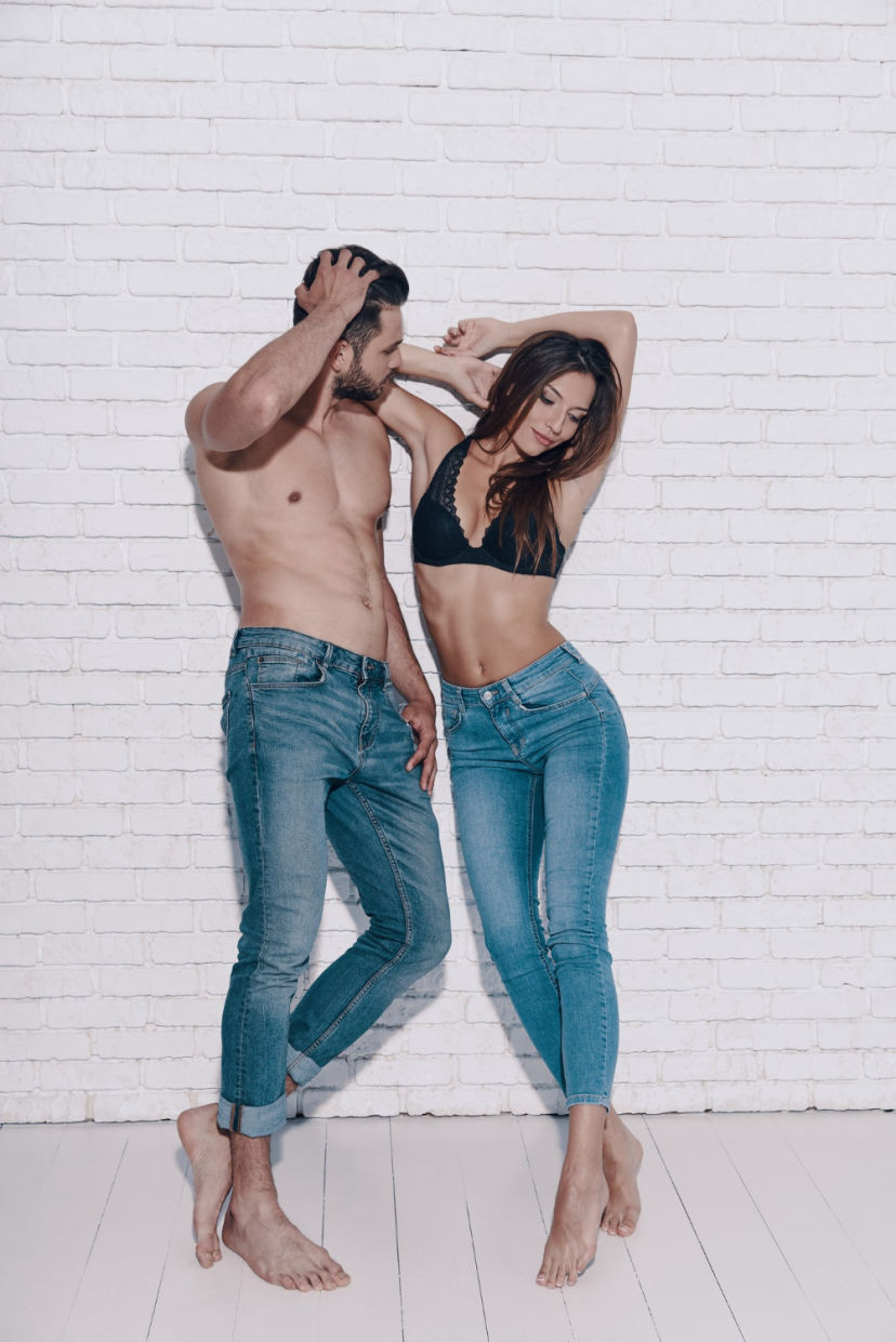 Hombre y mujer posando juntos frente a una pared de ladrillo blanco, ambos con vaqueros azules; él con el torso desnudo y ella con sujetador negro