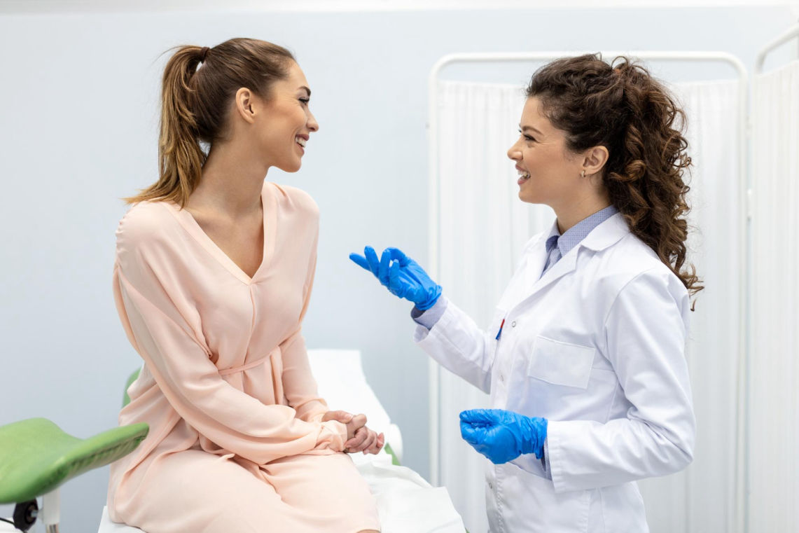 Mujer joven sentada con vestido rosa conversando con una doctora que lleva bata blanca y guantes azules